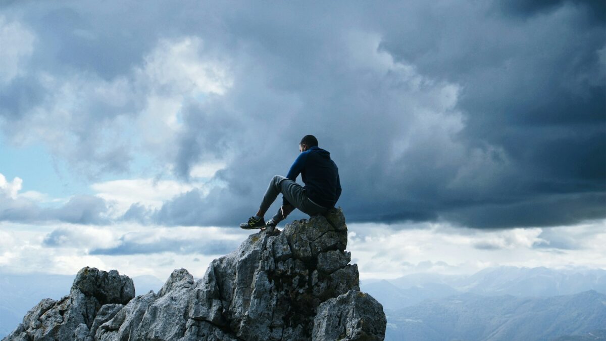 Man sitting atop a rugged mountain peak, contemplating a dramatic landscape under stormy skies.