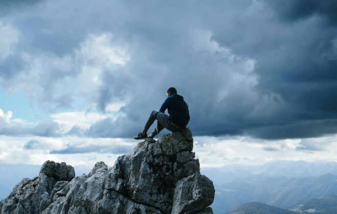 Man sitting atop a rugged mountain peak, contemplating a dramatic landscape under stormy skies.